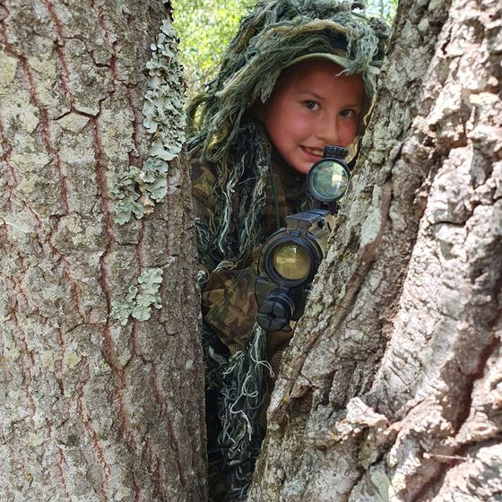A boy in a ghillie suit peeks around a tree
