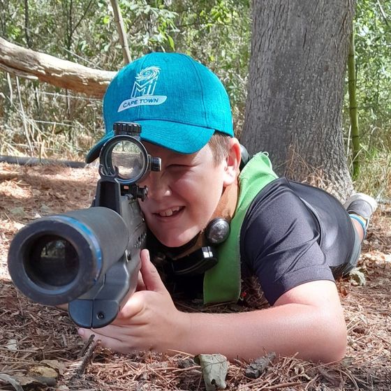 A boy in a teal cap aims while lying prone